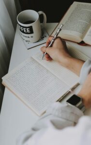 Person writing in a notebook on a desk with a coffee mug, creating a cozy workspace vibe.